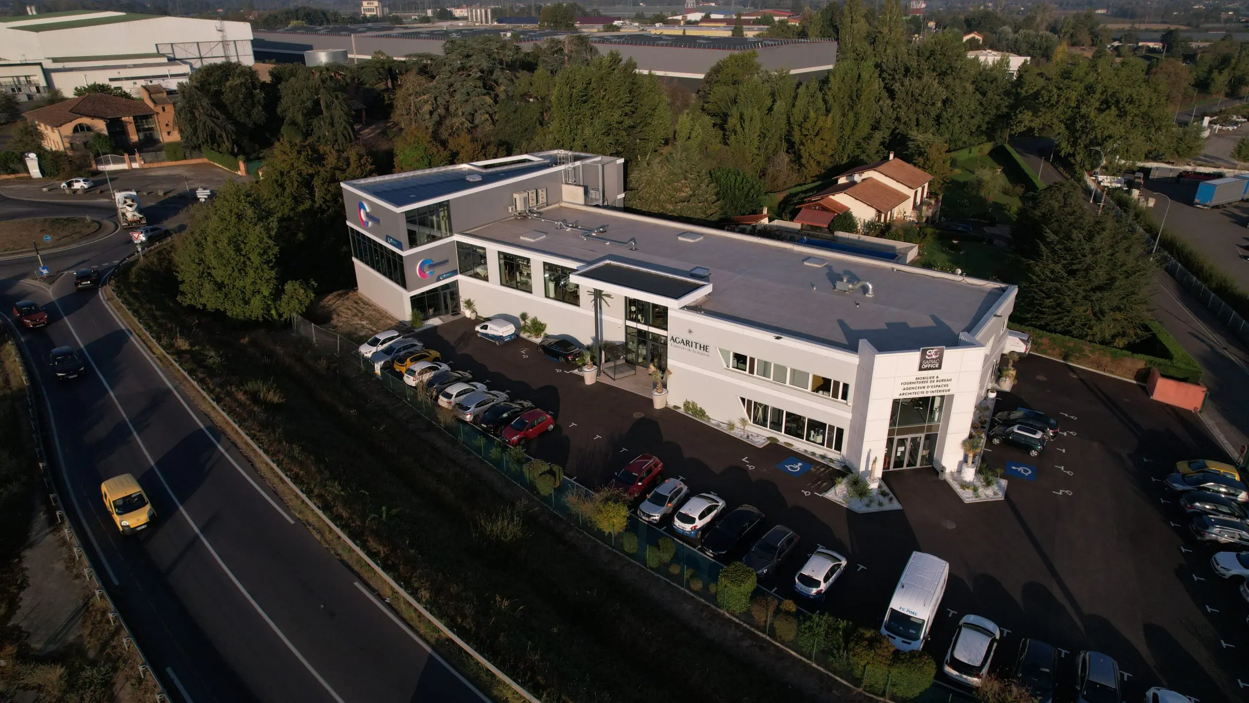 Vue aérienne d'un bâtiment moderne avec parking, entouré de verdure et situé près d'une route. Le bâtiment porte le nom "Novapage" et héberge plusieurs entreprises, avec des voitures garées à l'avant et sur les côtés.